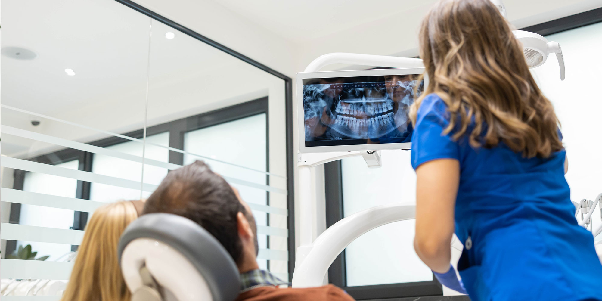 A dentist in blue scrubs shows dental X-rays on a screen to a patient seated in a dental chair
